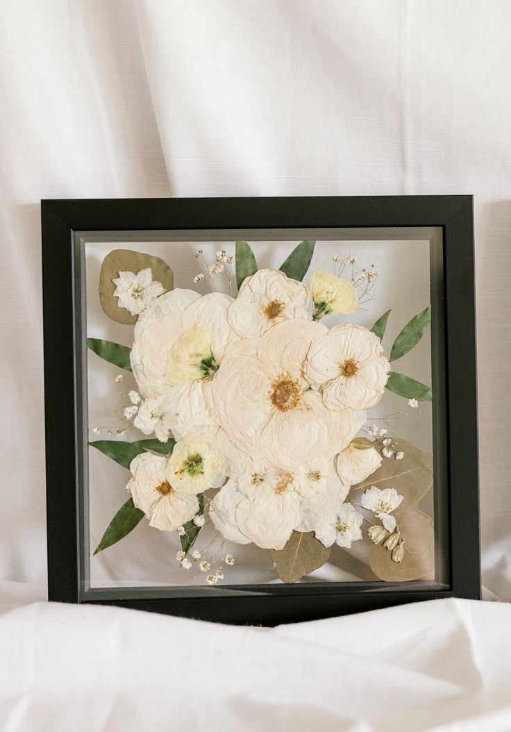 an arrangement of white flowers is framed in a black frame on a white bed sheet