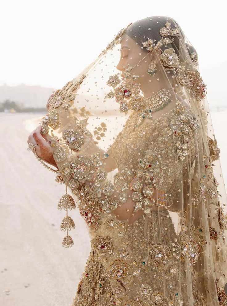 a woman in a gold wedding dress standing on the beach with her veil draped over her head