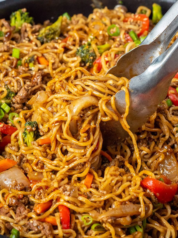 a skillet filled with noodles, meat and veggies being stirred by tongs