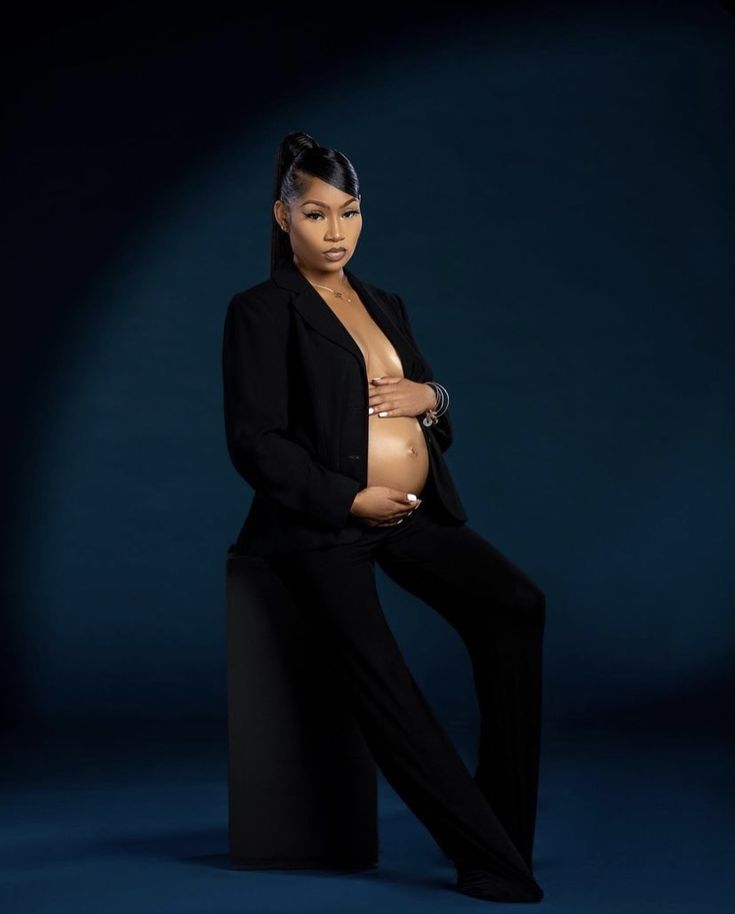 a pregnant woman sitting on top of a black stool with her arms around her stomach