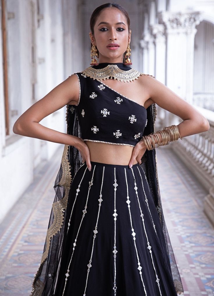 This look features a midnight black lehenga set. The lehenga has lines of gota and mirror work bootis descending into sequin & zari embroidered tiered borders. Teamed with a one shoulder blouse with pearl and mirror embellishments. A net dupatta with matching bootis and scallop border completes the look. Cancan & pockets are included. Composition: Blouse - Viscose georgette, lehenga -Viscose georgette ,Dupatta - Soft net Care: Dry Clean Only and Vacuum Storage This product can be customised for colour, sleeves, length of blouse and neck design Delivery : 2 weeks as the product is hand crafted. For more information and sizes please contact fabiliciousfashion@gmail.com or visit our Copenhagen studio.About the Designer : Renee Label was created in 2013,by the talented sister duo Pooja and Kar Black Mirror Work Dress, Black Mirror Work Lehenga, Lehnga Ideas, Shaadi Outfits, Black Dress With Pearls, Mirror Work Dress, Sequin Lehenga, Indian Bridesmaid Dresses, Mirror Work Lehenga