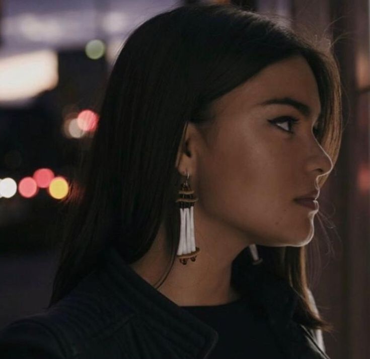 a woman with long hair wearing earrings and looking off into the distance at night time