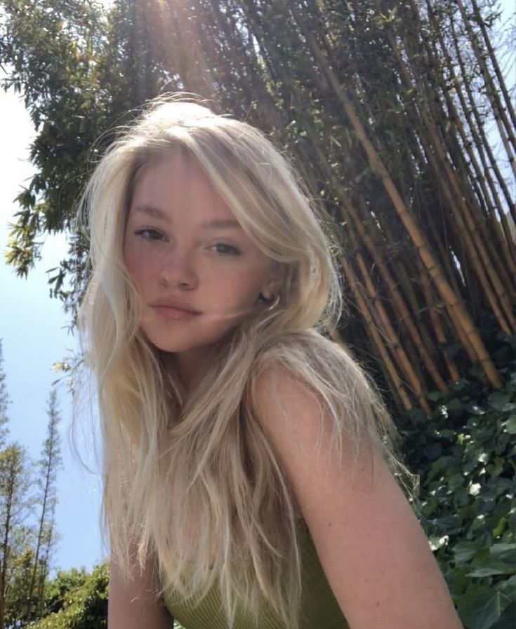 a woman with long blonde hair sitting in front of bamboo trees and looking at the camera