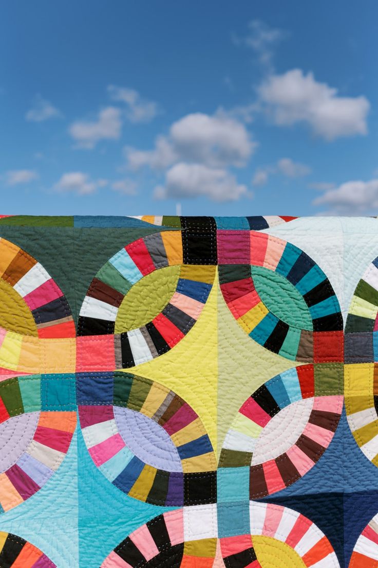 a colorful quilt on display in front of a blue sky with white clouds and bright colors