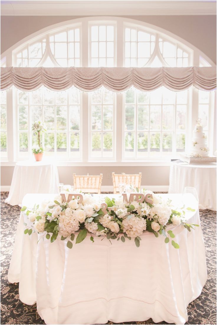 a table with white flowers and greenery on it in front of a large window