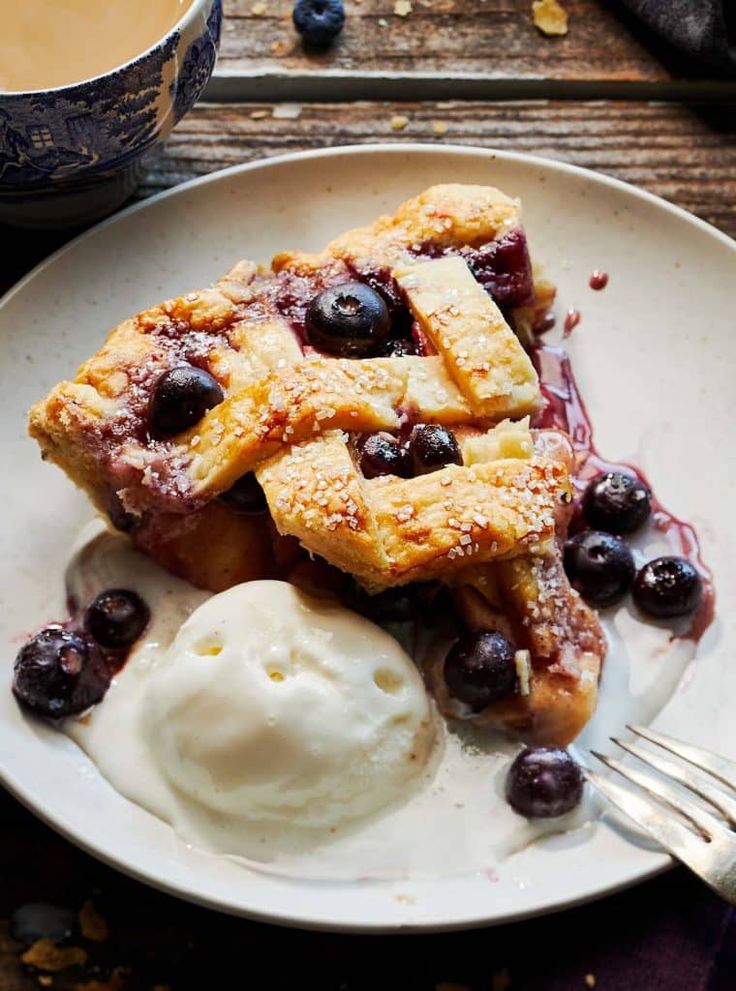 a piece of pie with blueberries and ice cream on a plate next to a cup of coffee