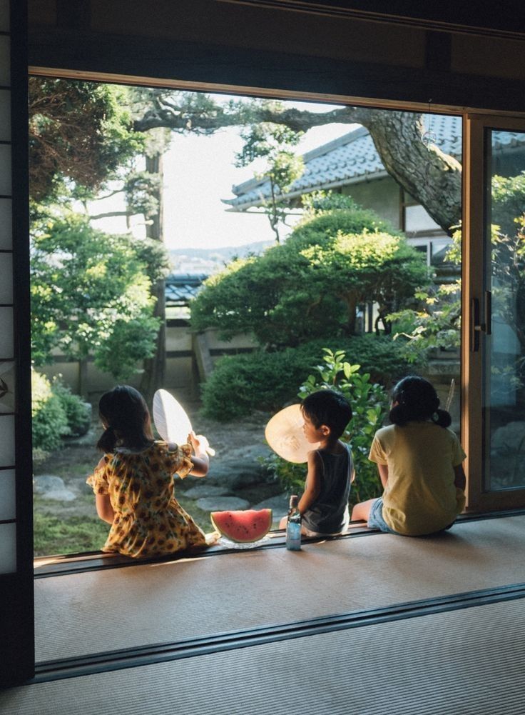 three children sitting on the windowsill looking out at an outside area with trees and bushes