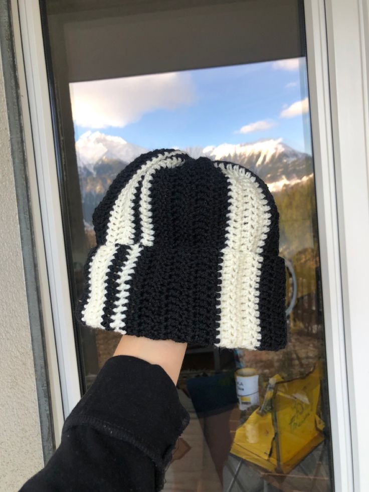 a hand is holding up a crocheted hat in front of a door with mountains in the background