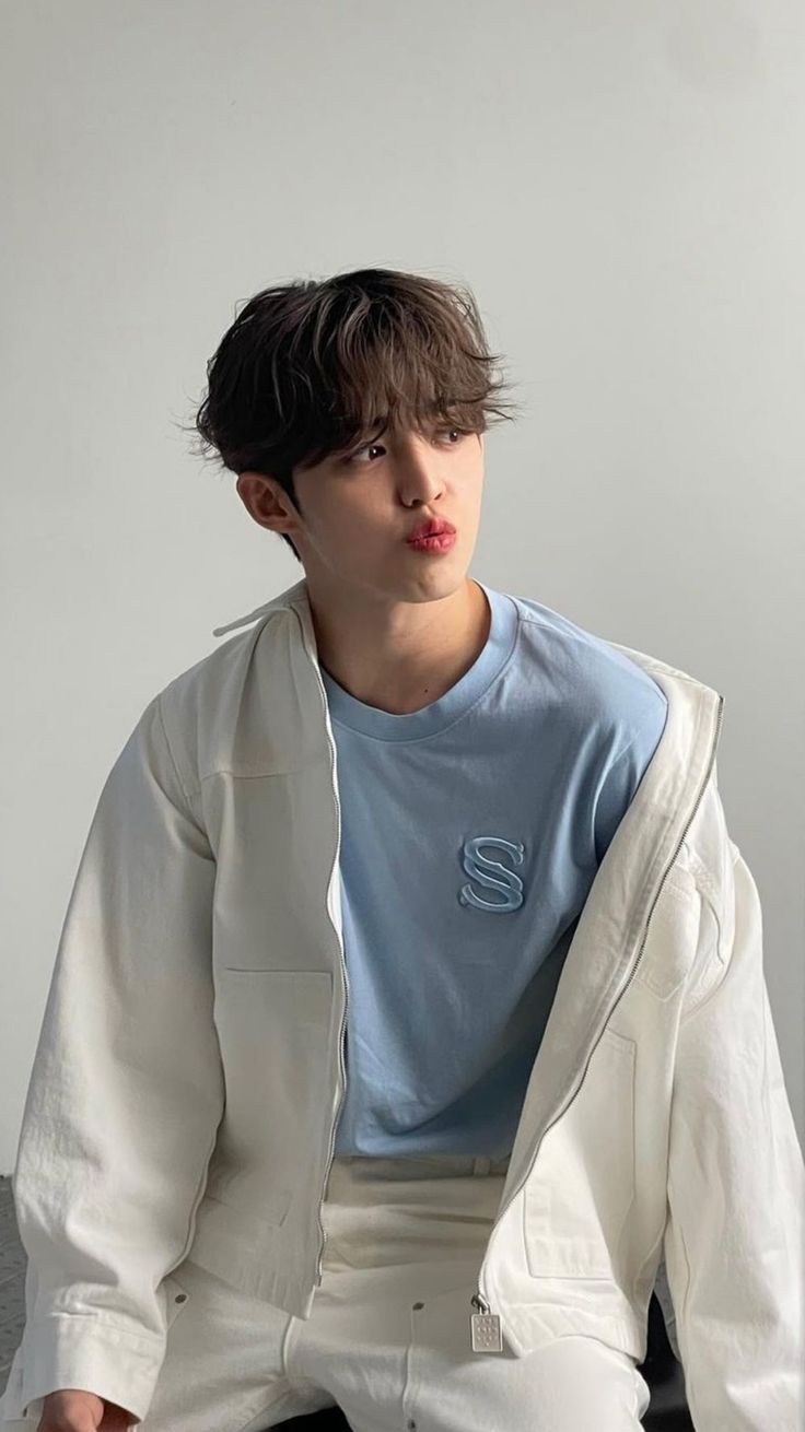 a young man sitting on top of a bed wearing a white jacket and blue shirt