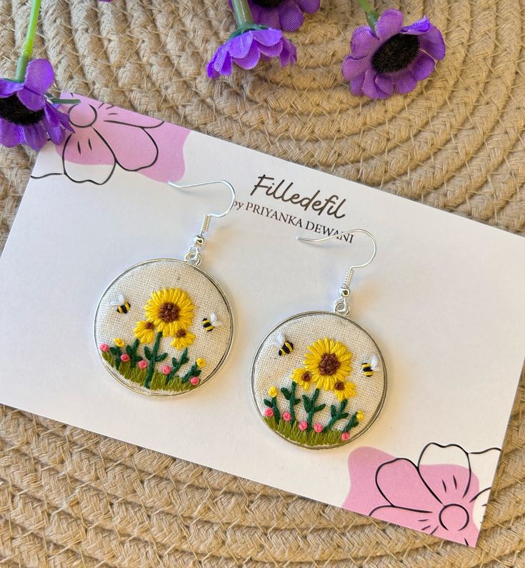 a pair of dandelion earrings sitting on top of a piece of paper next to purple flowers