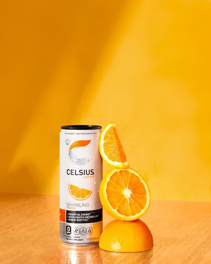 an orange is next to a can of celsius on a wooden table in front of a yellow background