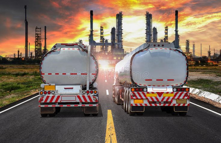 two tanker trucks driving down an empty road at sunset or sunrise with oil rigs in the background