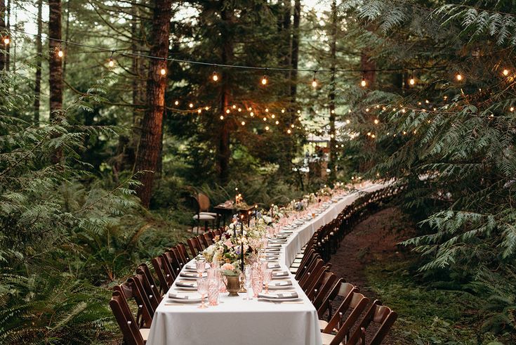 a long table is set up in the woods for an outdoor dinner party with string lights