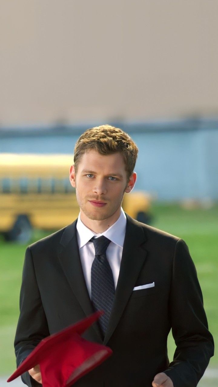 a man in a suit and tie is holding a red hat with one hand on his hip