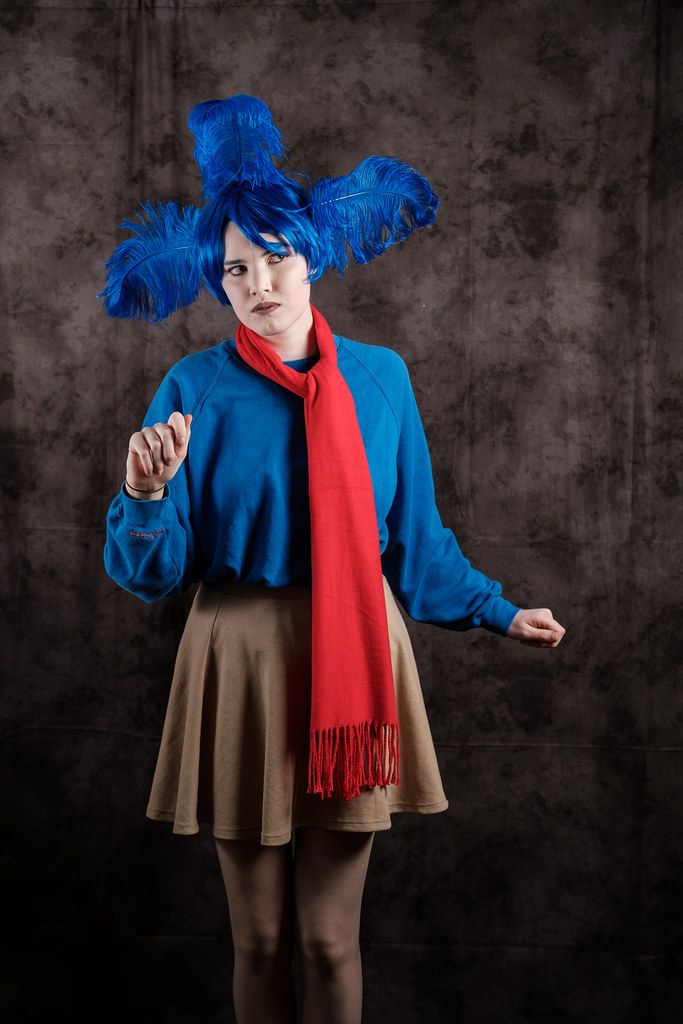 a woman with blue hair wearing a red scarf and brown skirt, standing in front of a dark background