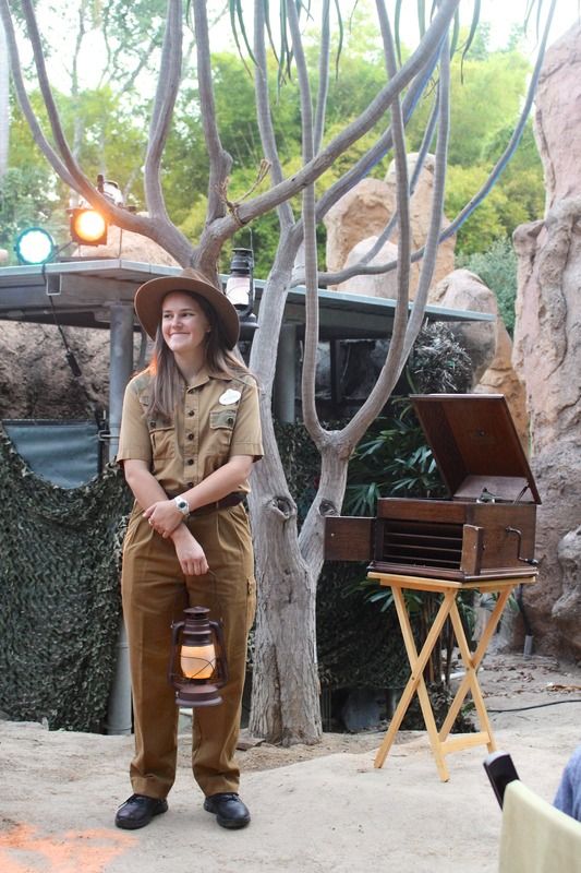 a woman wearing a hat and holding a lantern in front of an outdoor area with trees