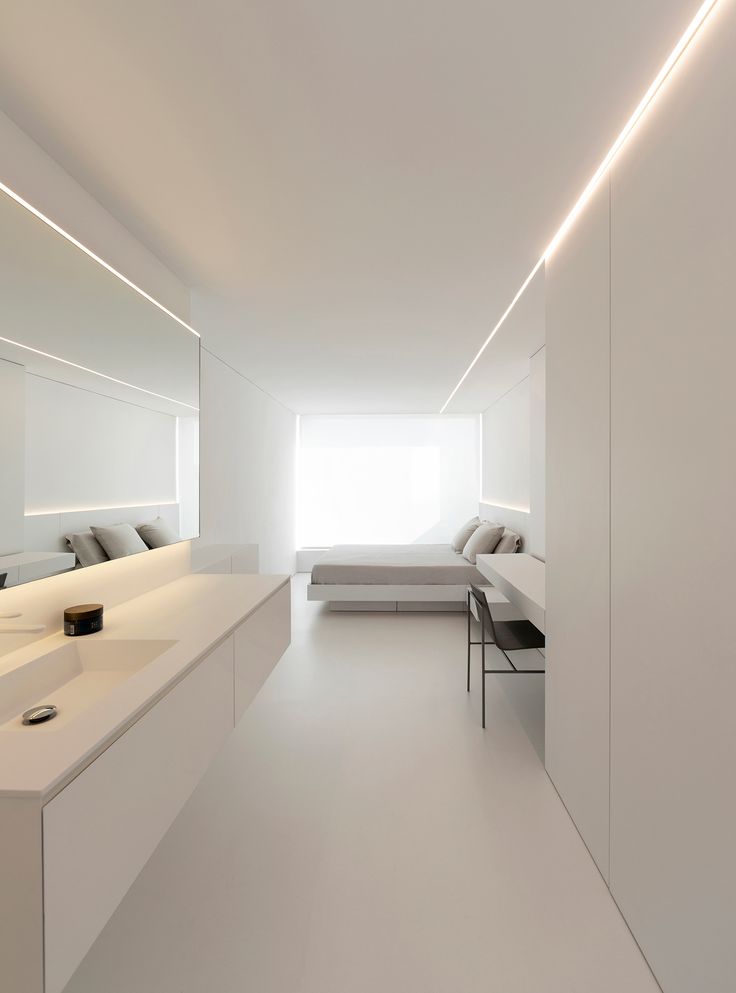 a long white room with a bed, desk and mirror on the wall next to it