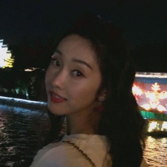 a woman standing in front of a river at night with lights on the water behind her