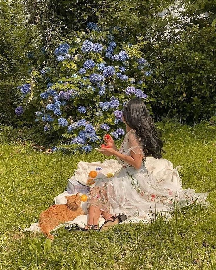 a woman sitting in the grass next to a cat and oranges on a blanket