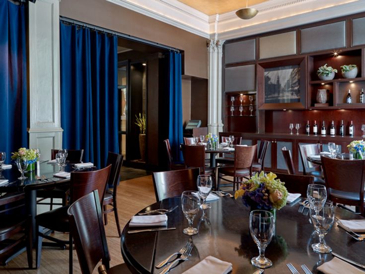 a dining room with blue curtains and wooden table set for four, along with wine glasses on the tables