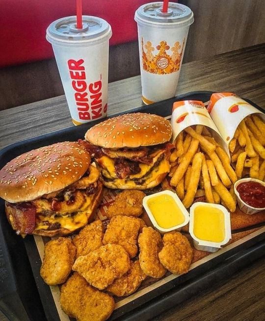 a tray with two burgers and fries on it next to cups of soda, ketchup, and condiments