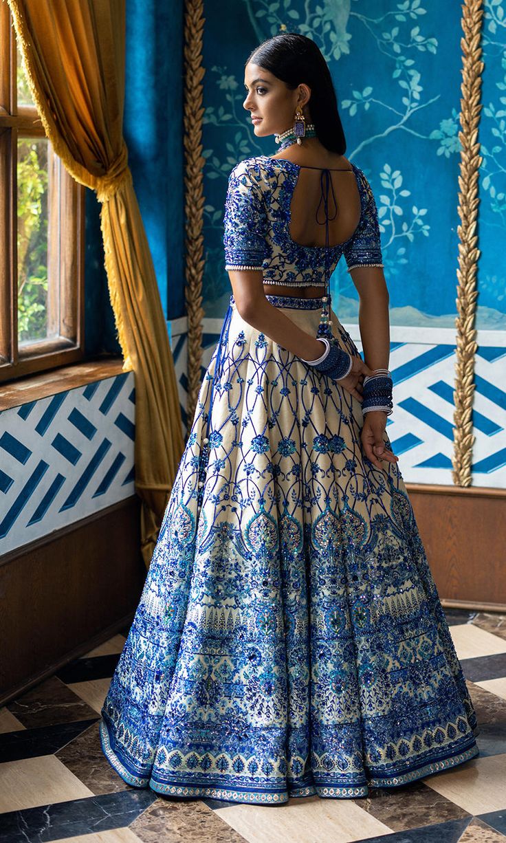 a woman in a blue and white dress standing by a window with an intricate pattern on it