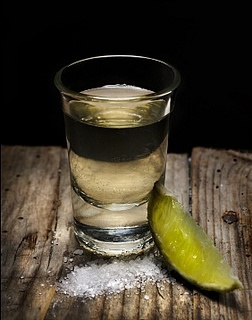 a glass filled with liquid next to a green pickle on top of a wooden table