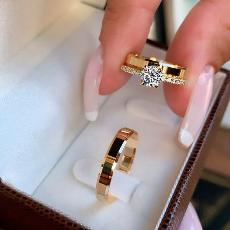 a woman's hand holding two gold rings in a box with the ring inside