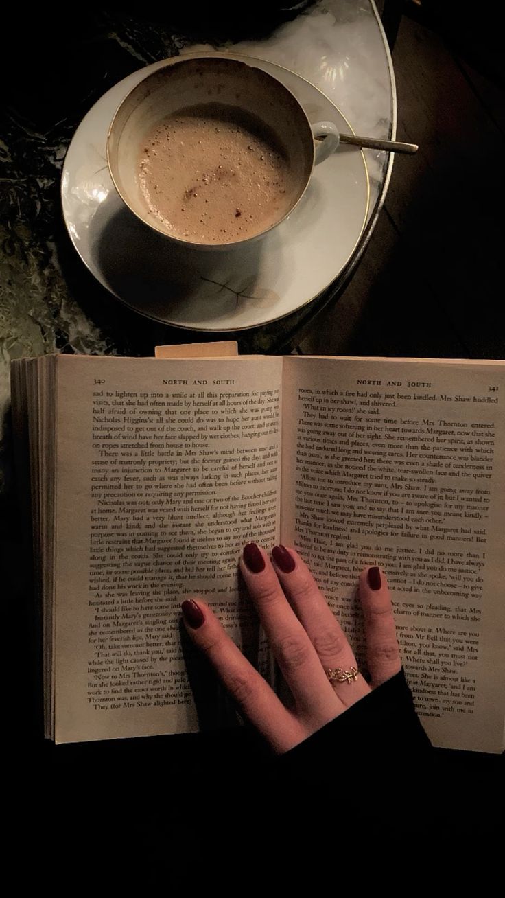 a woman's hand resting on an open book next to a cup of coffee