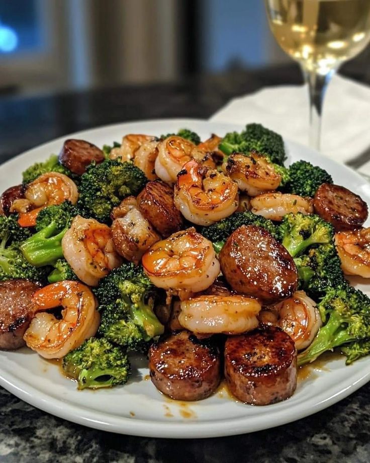 a white plate topped with shrimp and broccoli next to a glass of wine