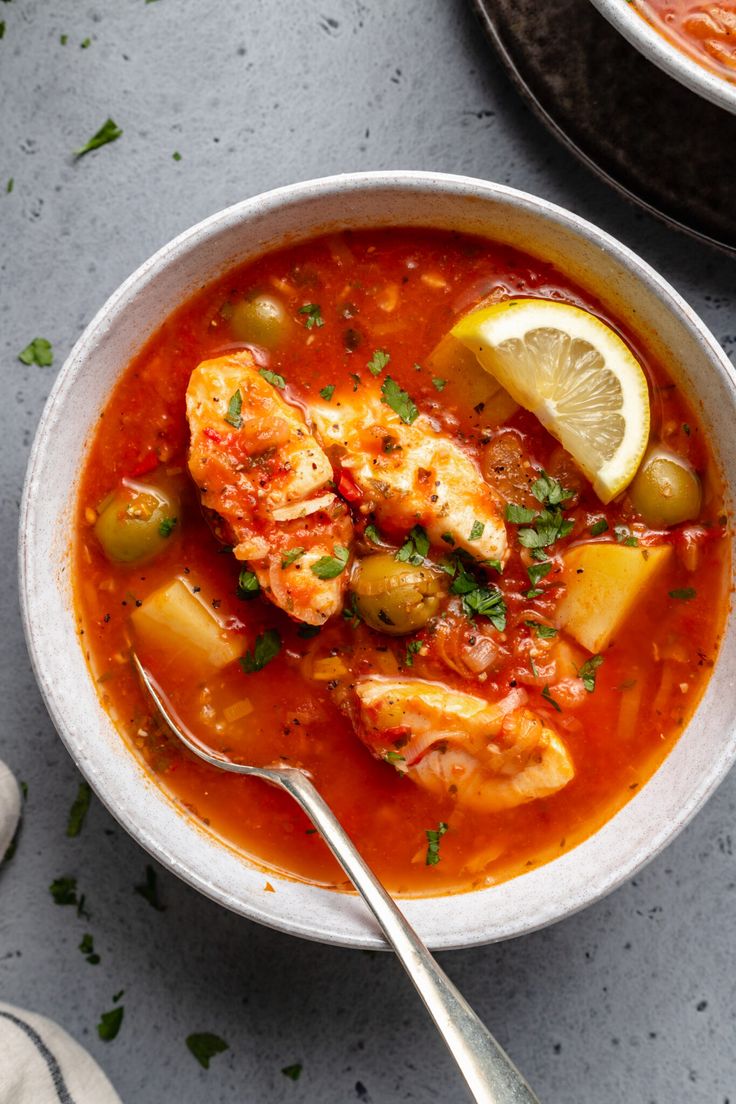 a bowl of soup with shrimp, olives and lemon in it next to a spoon