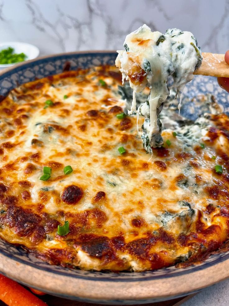 a person holding a spoon full of cheese and vegetables with carrots in the background