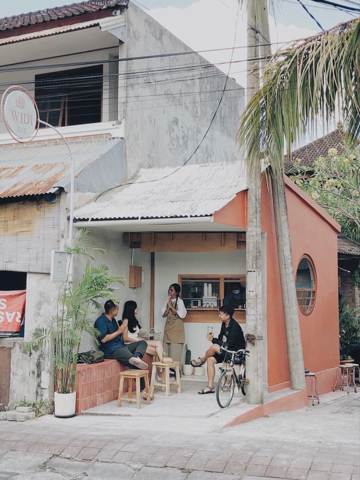 three people sitting at tables in front of a small building with palm trees on the sidewalk