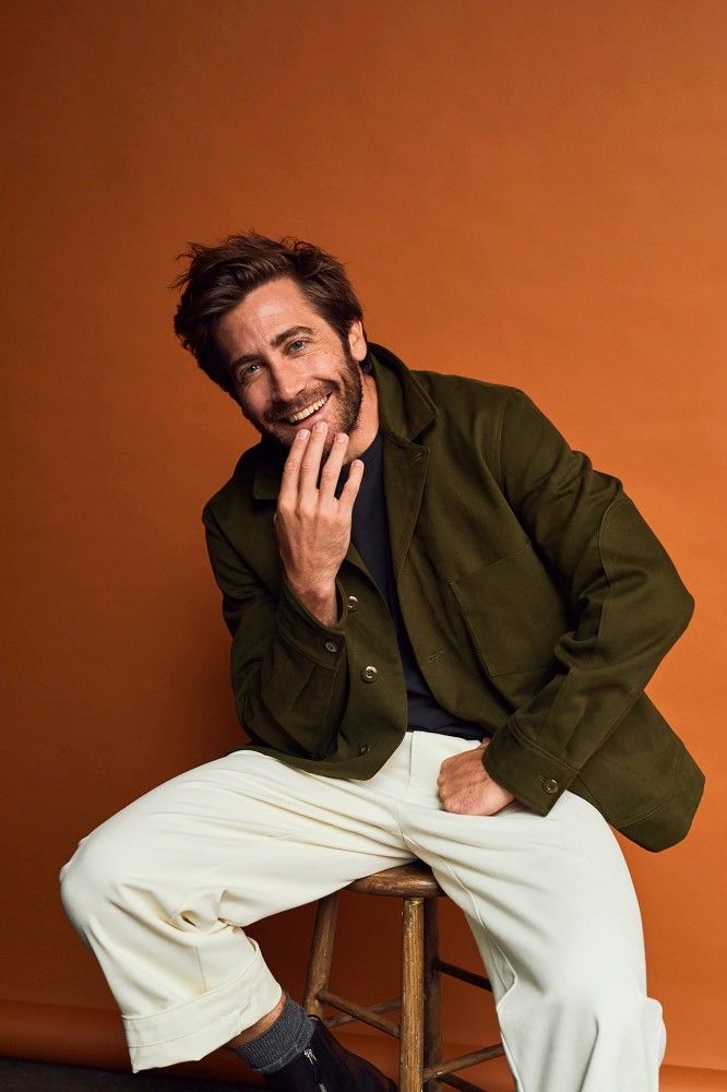 a man sitting on top of a stool next to an orange wall wearing white pants and a green jacket