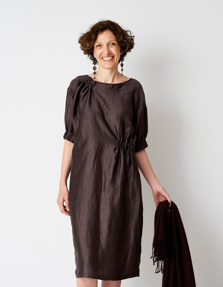 a woman standing in front of a white wall wearing a brown dress and holding an umbrella