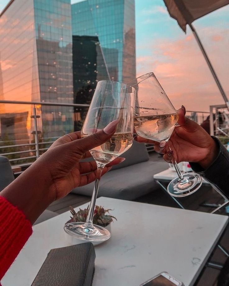 two people toasting with wine glasses on top of a roof at sunset in the city