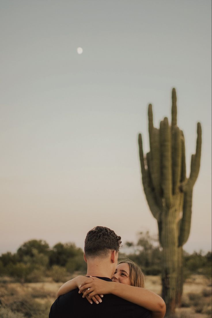 a man and woman hugging in front of a cactus with the moon in the background