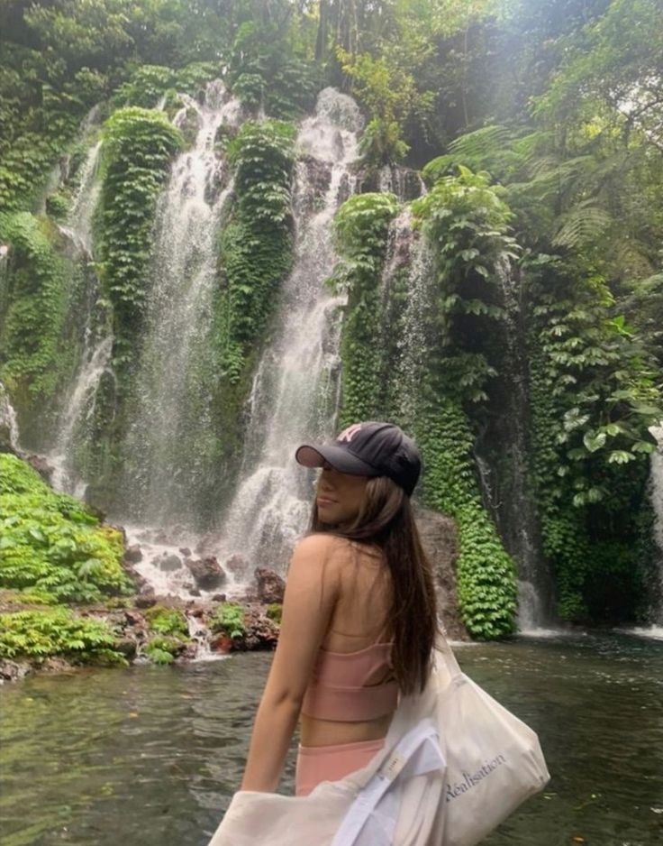 a woman standing in front of a waterfall with a bag over her shoulder and wearing a hat