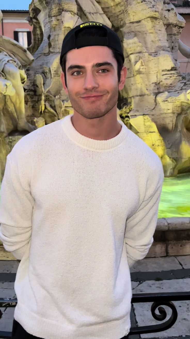 a man standing in front of a fountain wearing a white sweater and black hat with his hands in his pockets