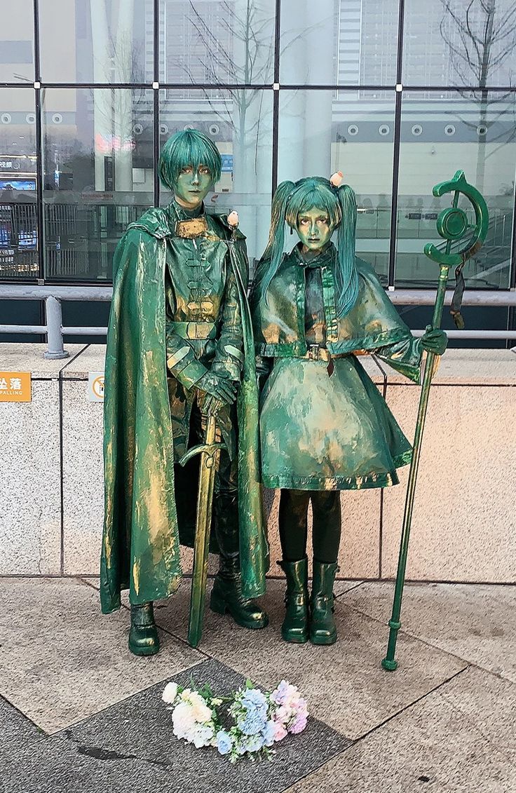 two statues of people dressed in green standing next to each other on the sidewalk near a building