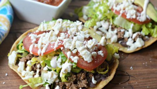 two tacos with lettuce, tomatoes and other toppings next to a bowl of salsa