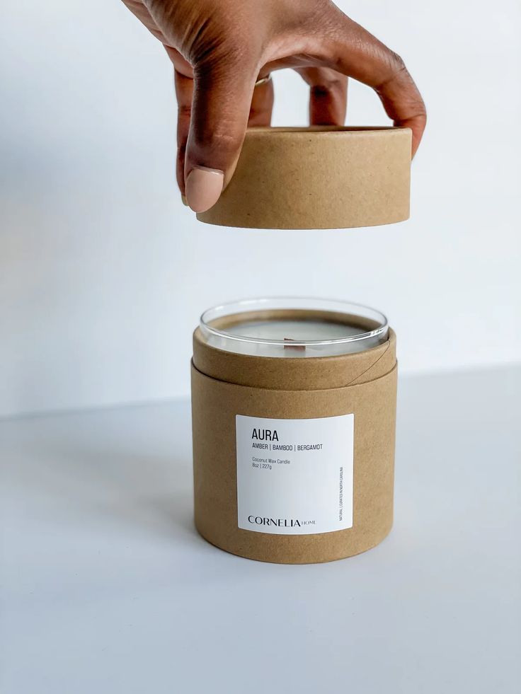 a hand is reaching for a candle in a brown container on a white table top