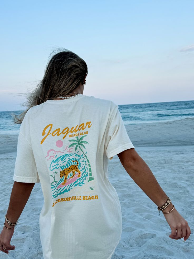 Our Sunhoney Jaguar Beach Club T-shirt is our newest exclusive design! This fun, funky design features bright pastel colors with a surfing Jaguar on the front and back catching a wave! This design is printed on Comfort Colors so you know it's going to be super comfy! Join the Jaguar Beach Club and rock this tee all summer long! Designed by us, handmade by us in Jacksonville Beach, FL Content: 100% Cotton Fit: TTS - size up for oversized fit Model wearing size: M Model sizing: 0-2 top, 24" bottom Fun Beach T-shirt With Sublimation Print, White Tropical Surfing Top, Casual Surfing T-shirt With Back Print, Tropical Crew Neck T-shirt With Screen Print, Graphic Print Vsco Tops For Summer, Graphic Print Vsco Summer Tops, Vsco Graphic Print Tops For Summer Adventures, Graphic Tee For Surfing With Sublimation Print, Beachy Crew Neck T-shirt For Surfing