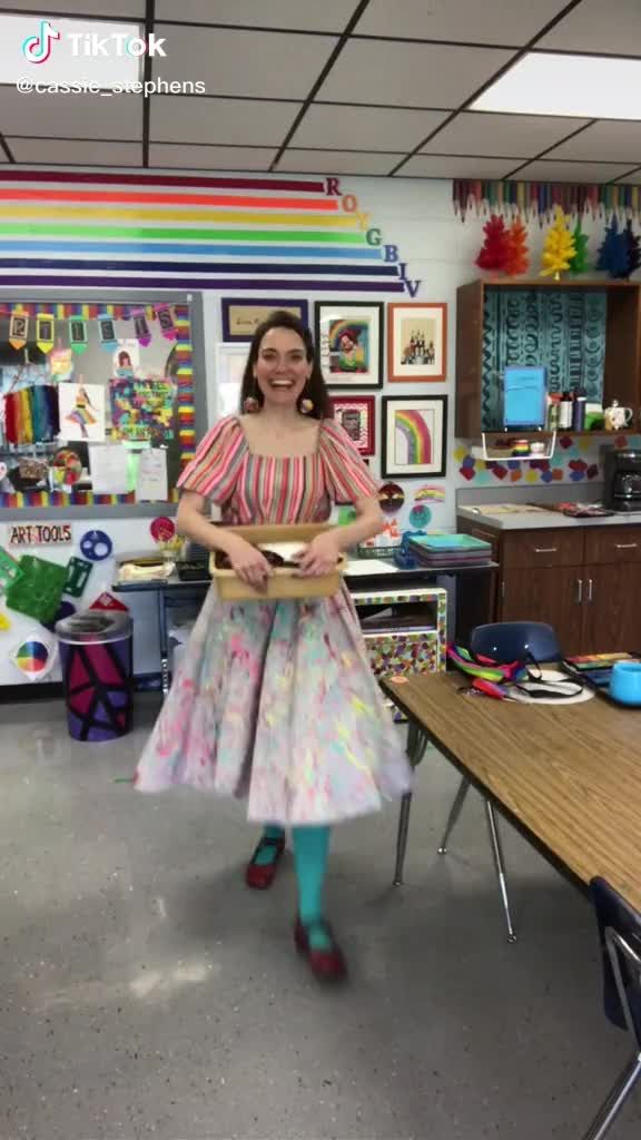 a woman in a colorful dress is holding a tray and smiling at the camera while standing next to a table