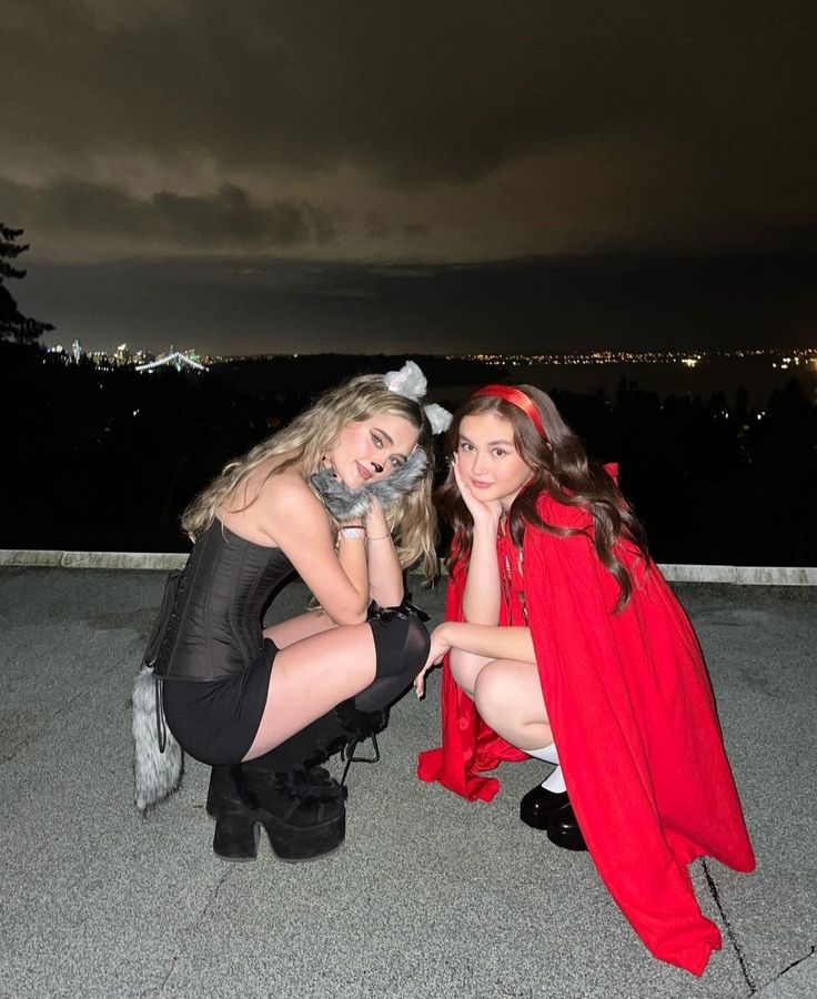 two beautiful young women dressed in costumes sitting on the ground at night with city lights behind them