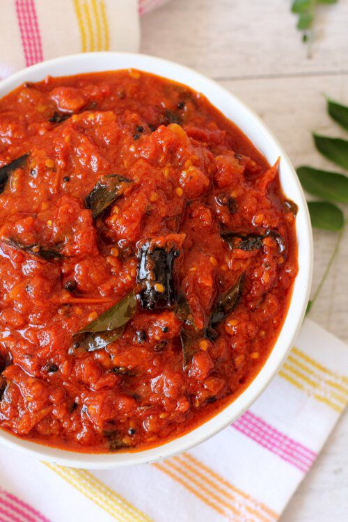 a white bowl filled with tomato pickle sauce on top of a colorful napkin and green leaves