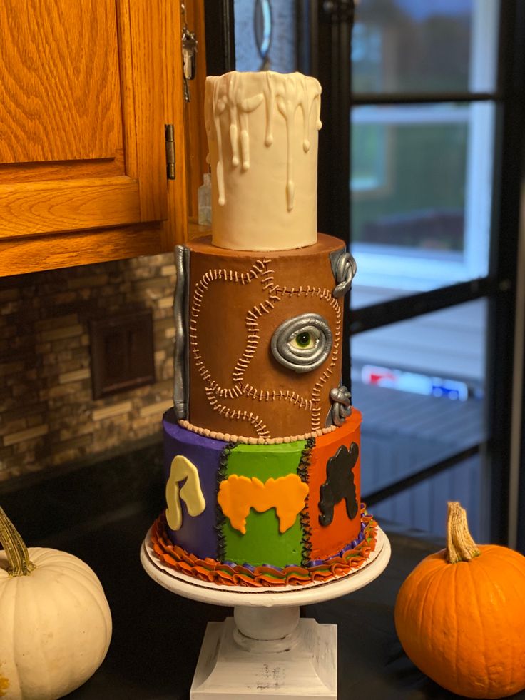 a multi - tiered cake decorated with colorful designs and decorations sits on a table next to two pumpkins