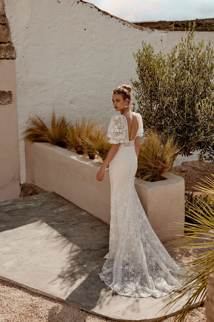 a woman standing in front of a white wall wearing a wedding dress with an open back