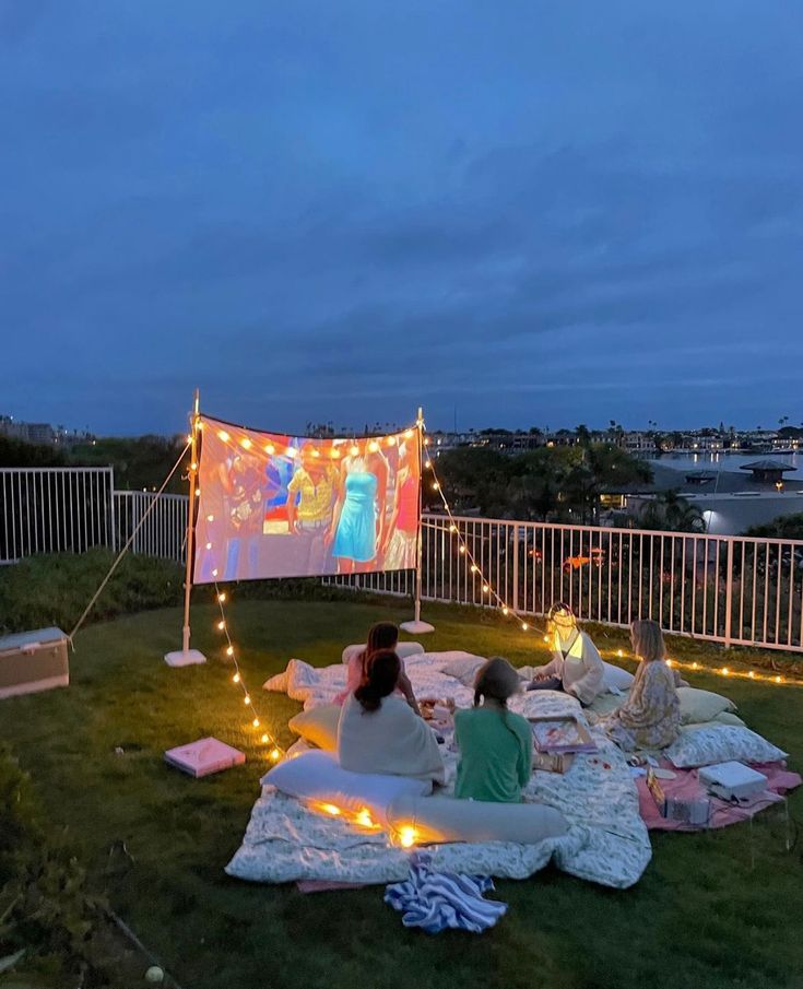 two women sitting on blankets in the grass with lights strung around them and an outdoor movie screen behind them