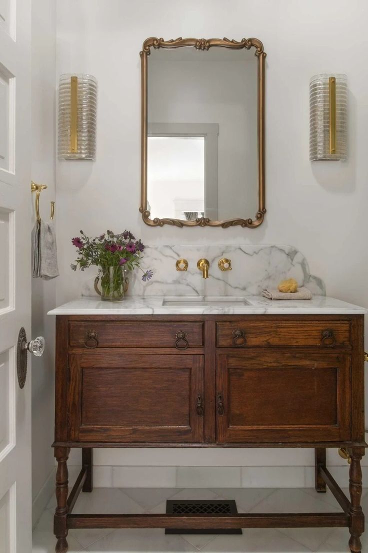 a bathroom vanity with marble top and gold accents on the mirror above it, along with two brass sconces